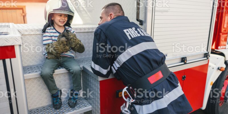 istockphoto-1186054054-2048x2048 Little boy playing with firefighter helmet and gloves and talking with real firefighter